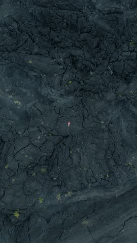 Aerial view of a person lying on dark volcanic rock, surrounded by cracks and sparse vegetation, highlighting the raw beauty of the volcanic landscape.
