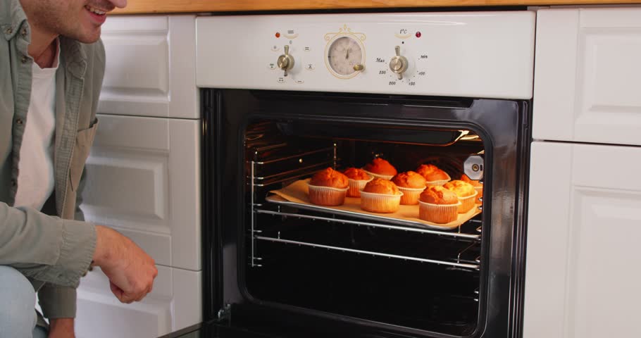 Happy man confectioner taking tasty muffins out of oven, looking with cheerful expression at delicious pastries ready for eating, demonstrating excellent culinary skills, being at home