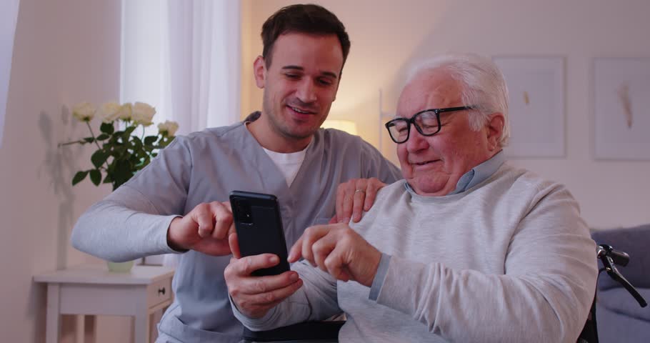 Young nurse showing elderly man in wheelchair how to use smartphone. Nursing home service and patient support. Caregiver teaching senior person to handle with mobile phone. 4k video.