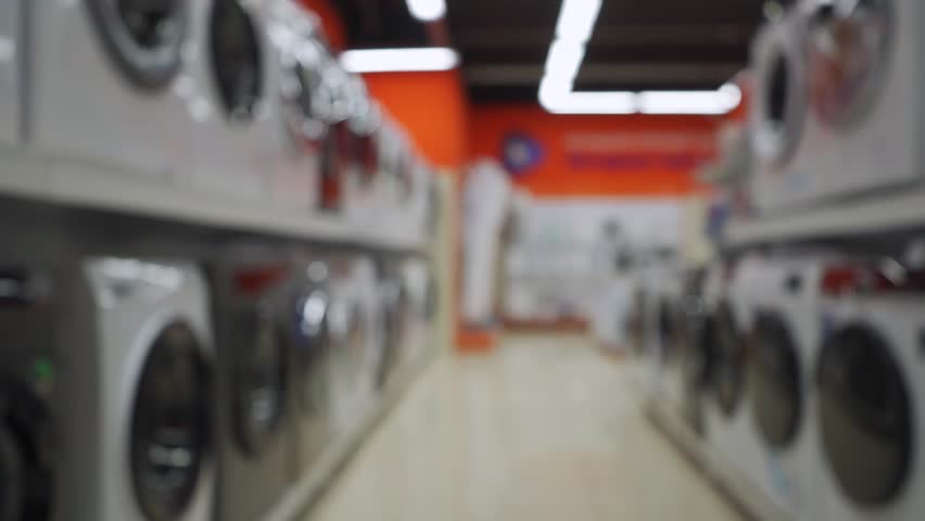 A blurred view of a row of washing machines displayed in a brightly lit appliance store, emphasizing a modern and spacious design.