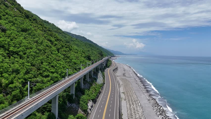 A train of EMU3000 series dashing on the elevated tracks of South-Link Railway parallel to a coast highway between green mountains and the azure Pacific Ocean near Duoliang Station in Taitung, Taiwan
