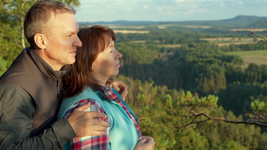  Happy middle-aged couple on top mountain in national park Saxony, Germany. Man shows his hand in distance at magnificent scenery: cliffs, forests