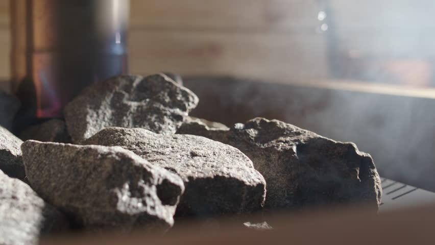 Water sizzling and steaming as copper ladle pours it over sauna rocks, closeup