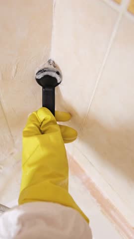 Cleaning tiles with a brush, a specialist in a white suit tackles mold on the bathroom walls using effective cleaning methods.