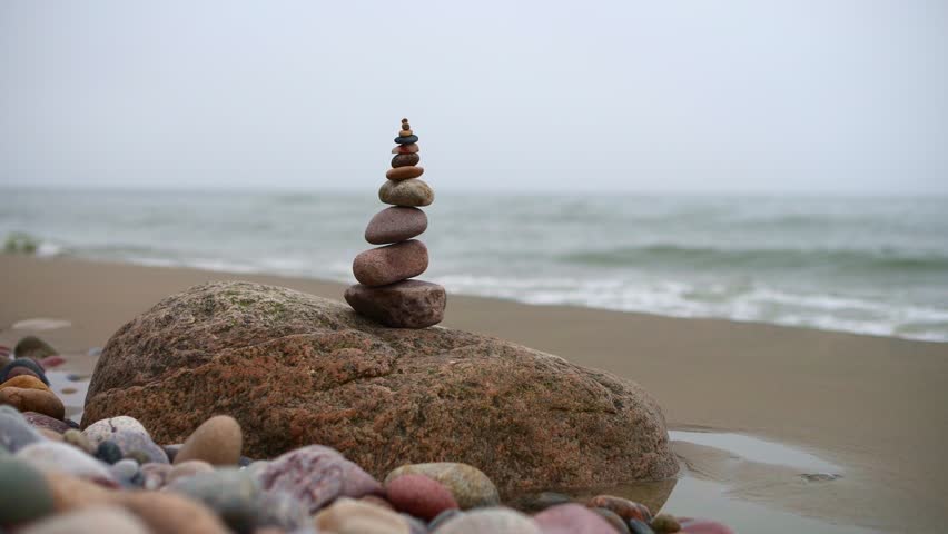 Balanced pebble stack on large rock by ocean. Overcast sky, serene beach scene with gentle waves. Foreground detail includes colorful pebbles, emphasizing tranquility