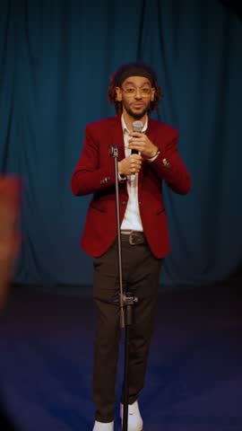 Full-length portrait of professional comedian on stage, vertical video with funny African American man. People applauding to black guy with microphone, new stand-up comedy show in bar or nightclub
