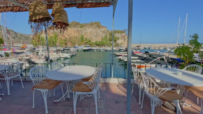 Slowly passing an outdoor harbour restaurant in Marina del Este, Andalusia, Spain. Hanging lamps sway gently in the breeze. Motorboats and sailboats are moored in the background, enhancing the scene.