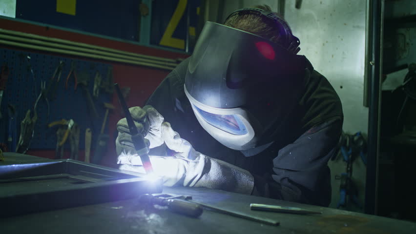 Welder in protective workwear, helmet, gloves, and coverall working in a workshop, welding metal bars, close up shot. Craft, skill, and manufacturing concepts.