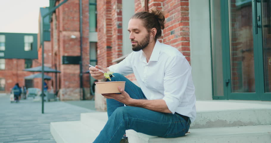 Business man in white shirt eating healthy salad on a break sitting on stairs over office building background. Male dieting nutrition concept