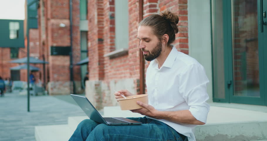 Man eating takeaway healthy salad outdoors and watching videos on laptop relaxing on stairs in front of office building on street