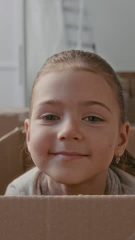 Vertical shot of adorable little girl looking at camera while peeking out of big cardboard box in domestic room of new apartment during relocation or home renovation