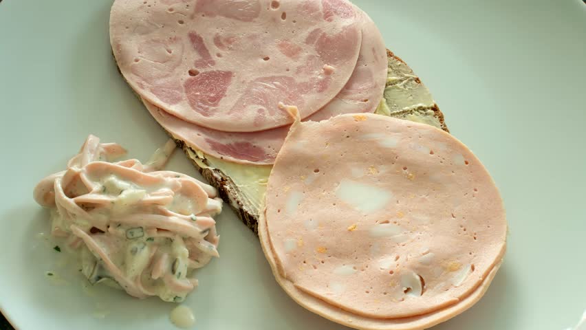 Zoom on buttered slice of brown bread on a white plate. Two slices of ham sausage and two slices of egg sausage on the bread. Besides sausage salad with mayonnaise.