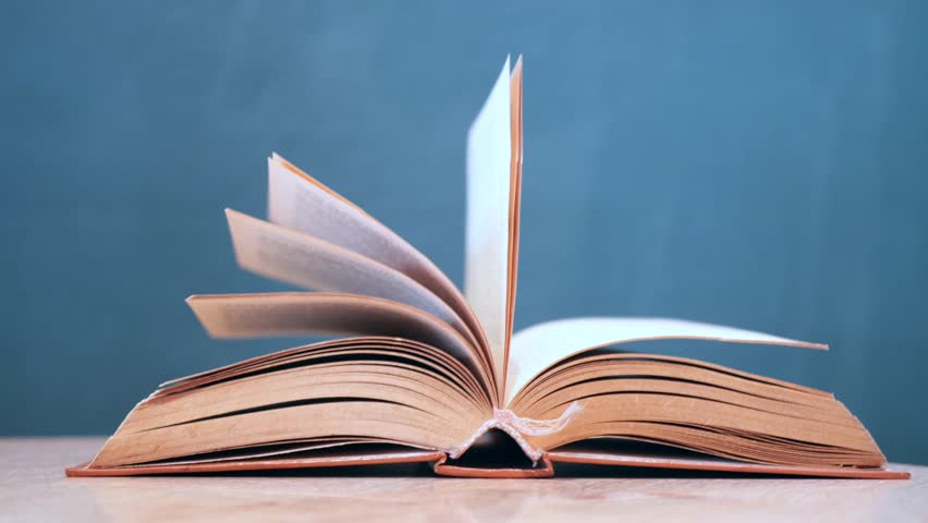 An open book with its pages gently turning rests on a wooden table in the breeze