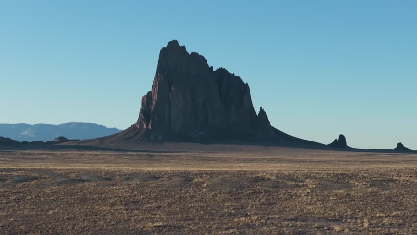 A low to the ground, slow-rising 70mm drone shot of the sacred “Ship Rock”, a massive, monolithic monadnock towering above the high-desert plain of the Navajo Nation, in New Mexico, United States.