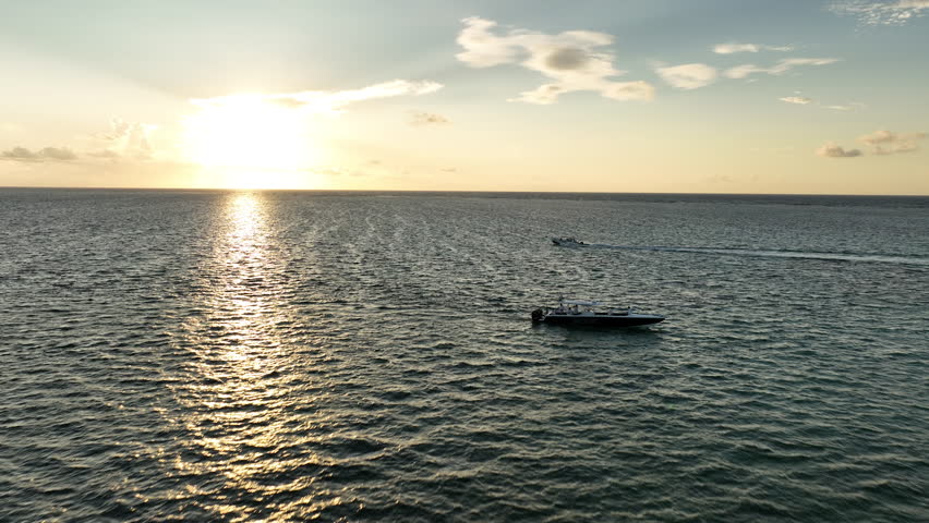 A breathtaking drone shot captures a boat speeding across the Caribbean ocean at golden hour sunrise. The warm sunlight reflects off the tranquil waters, with a stunning coastline in the background.