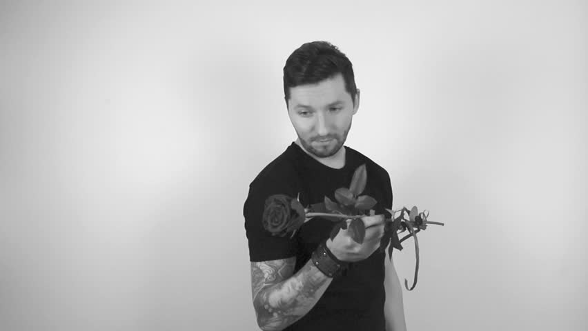 Grayscale footage of a tattooed man with short hair and trimmed beard looking at a blossoming rose flower, smelling it and offering in front of him to the camera or the viewer in slow motion.