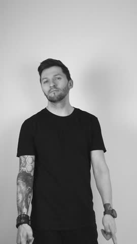 Grayscale footage of a man with tattoos short beard and hair lifting up his arms and pointing straight forward at the camera with a confident face and smile indicating he approves and agrees with you.