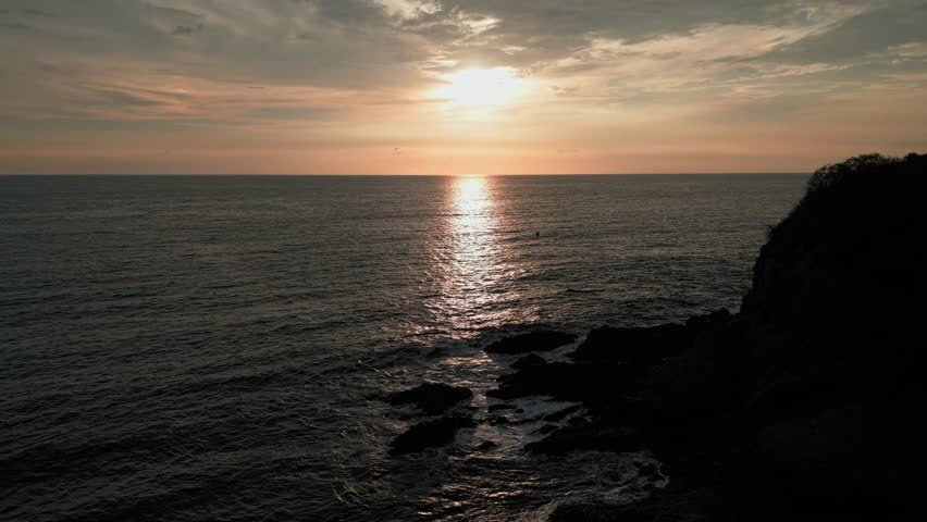 Aerial 4K shot over the ocean at sunset near Sabalo Beach and Hill (Cerro del Sabalo) in Mazatlan. The camera moves forward towards the horizon, with the sun reflecting on the water and light waves.