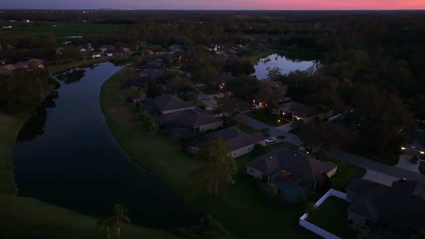 Luxury villas and homes in Florida at dusk scene. Aerial rising wide shot. Private lake in Plant City neighborhood. Sunset time at horizon with colored sky. Establishing shot.