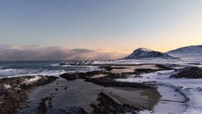 Aerial drone gliding and flying over black rock, sand and snow covered shoreline in a beach at Norway during winter time with mountains in the background during sunset or sunrise with sky orange color - Powered by Shutterstock - Get 15% off with code: PIKWIZARD15