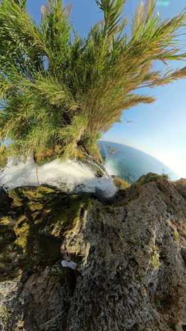 Vertical video. A breathtaking view of water cascading down a cliff, merging into the vast, blue sea, captured from a high-angle, wide perspective, highlighting the rugged cliffside and expansive ocea