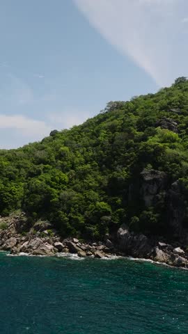 Rocky shoreline framed by lush green hills and deep blue ocean under a clear sky. Koh Tao, Thailand.