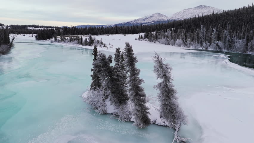 An aerial establishing shot captures the frozen Lake Kusawa and Takhini River winding through snow-covered fir forests with mountains on the horizon, epitomizing a pristine Yukon winter wilderness.