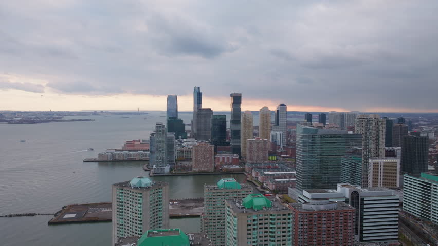Jersey City skyline glimmering at sunset, clouds hovering over Hudson River, urban landscape in soft evening luminescence