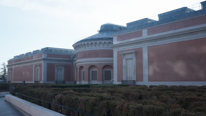 Prado Museum in Madrid, a large red brick structure featuring a rounded section and flat roofs, with trimmed shrubs in front, located in Spain’s capital city