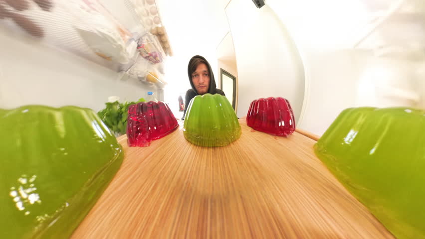 From inside the refrigerator, a man places a tray of wobbly red and green jelly, closing the door as the soft-textured jellies jiggle slightly on the shelf
