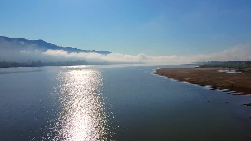 Aerial view of a tranquil river reflecting sunlight, with a scenic backdrop of misty mountains and a sandy shoreline. The serene atmosphere captures the beauty of nature in the early morning