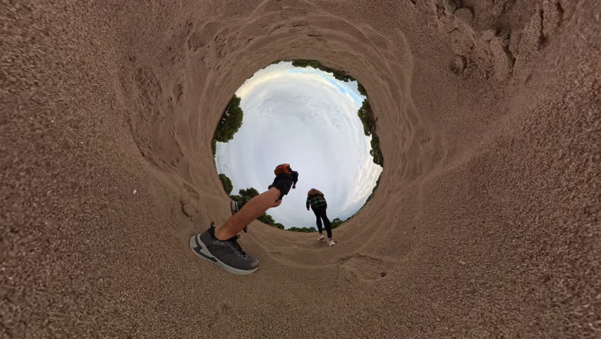 A slow-motion scene shows a man and a woman traveling on foot in a desert-like area, running uphill on a sandy path. Their steps kick up sand, framed by the natural surroundings of sparse greenery