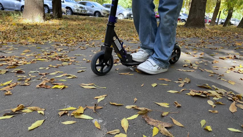 Close up of person riding a scooter on an autumn park path covered with fallen yellow leaves