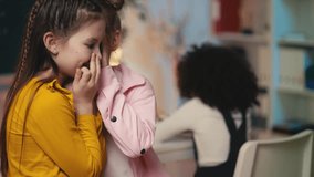 Two little girls gossiping about a classmate in the classroom, bullying concept - Powered by Shutterstock - Get 15% off with code: PIKWIZARD15