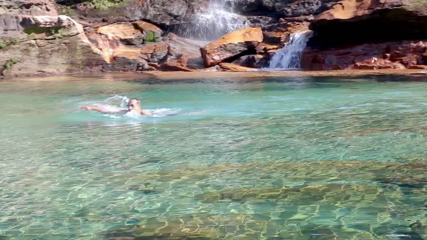 young man jumping and swimming in blue clear water of waterfall from flat angle
