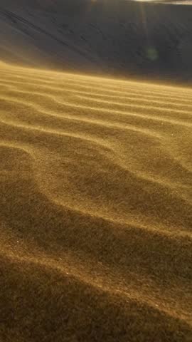 Grains of sand waving in the wind. Golden sand dunes create mesmerizing patterns in early morning light at remote desert location with undulating terrain. Slow motion vertical shot
