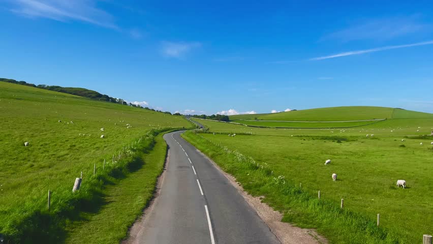English countryside road stretches through vibrant green hills under a bright blue sky. The peaceful scenery embodies the charm of rural tranquillity and timeless natural beauty
