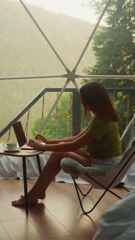 Woman works on laptop on vacation in bright room of glamping house. Young woman alone sits in armchair and types text on keyboard with cup of coffee on table