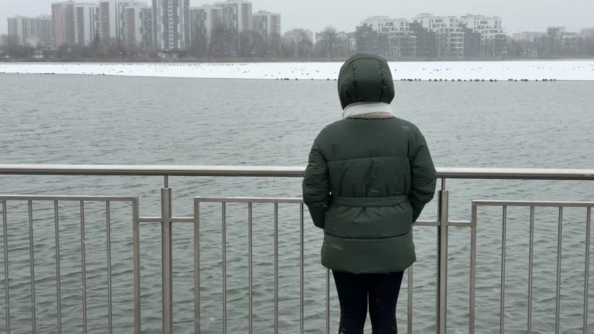 Young caucasian woman walks near a city lake on a snowy winter day and looking at birds. Ivano-Frankivsk, Ukraine
