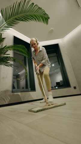Middle-aged woman having fun while mopping the floor and listening to music with headphones in a modern room with a potted palm. High quality 4k footage