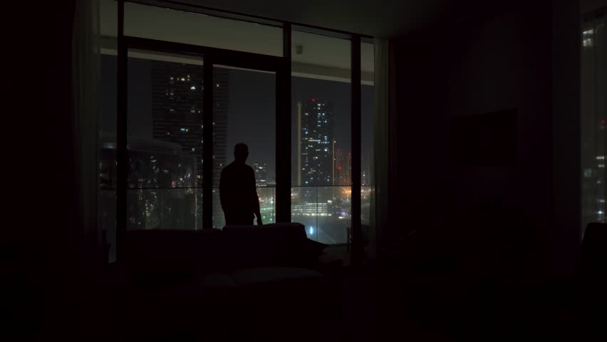 Silhouette of a man with insomnia, loneliness, or depression in an apartment overlooking a city at nighttime