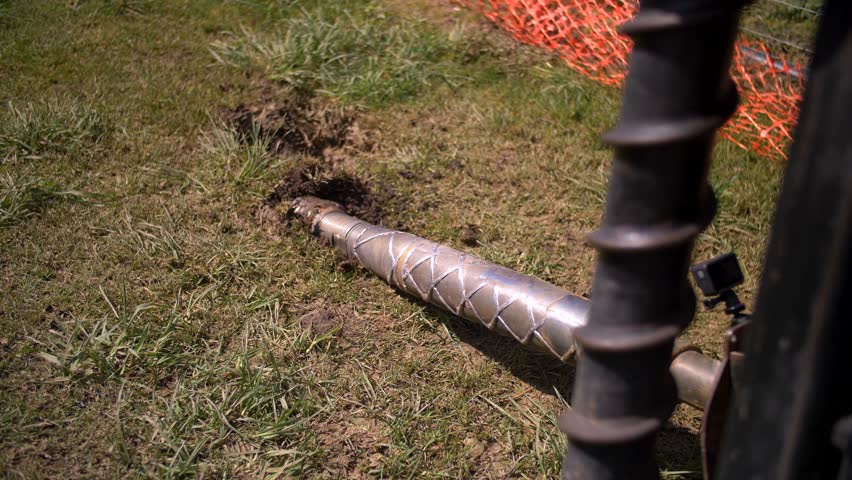 A steel drill head emerging from the soil, highlighting its spiral design for underground boring. The muddy surface and disturbed earth show recent excavation work for pipeline or utility installation