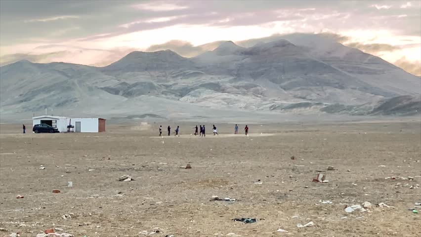 Footage of unrecognisable bunch of kids playing soccer in desert by sunset. Dry coast of Peru with mountains in background, unique environment