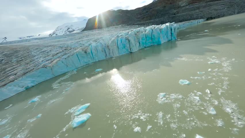 FPV aerial drone flying close to icy blue glaciers and in between them on a sunny day between Greenland or Iceland mountains that are covered with some snow. The Glaciers are in brown ocean water.