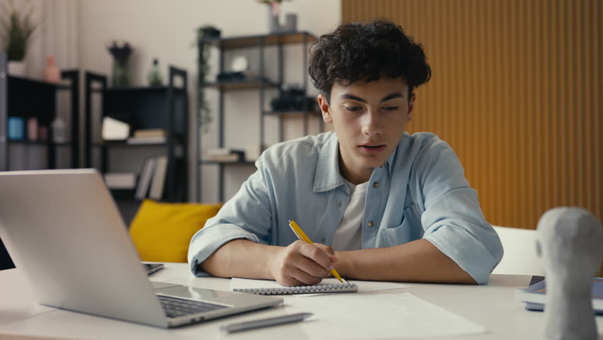 Teenager studying at home, writing in a notebook and using a laptop for research