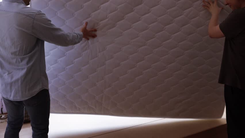 A couple carry a mattress into the new apartment. Laid and covered the new mattress on the slat of bed in bedroom.