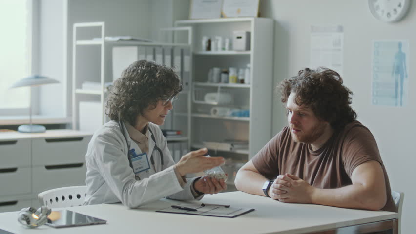 Female doctor in white coat discussing diet plan with obese patient, giving him bottle of pills and explaining dosage during medical consultation in clinic