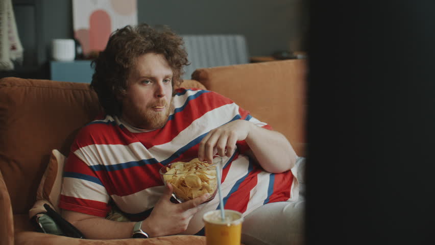 Obese man resting on couch, eating chips from bowl and drinking soda as watching TV in cozy living room during evening at home