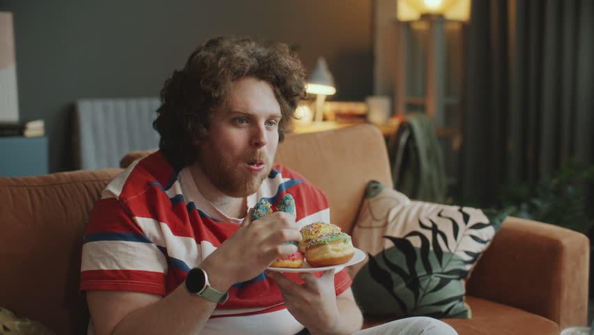 Young obese man sitting on sofa in living room, holding plate of sweet glazed donuts and eating them while watching TV show in evening at home