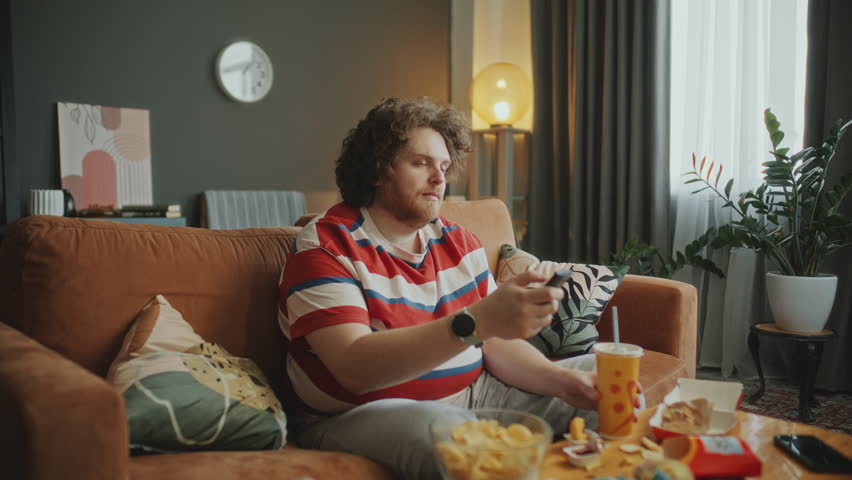 Obese man turning on TV with remote control, watching programme, eating snacks and drinking soda while sitting on couch near table filled with fast food in living room. Zoom shot